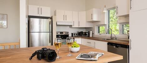 A modern kitchen with white cabinets, stainless steel appliances, a wooden island with a camera, wine glasses, and a bowl of fruit.