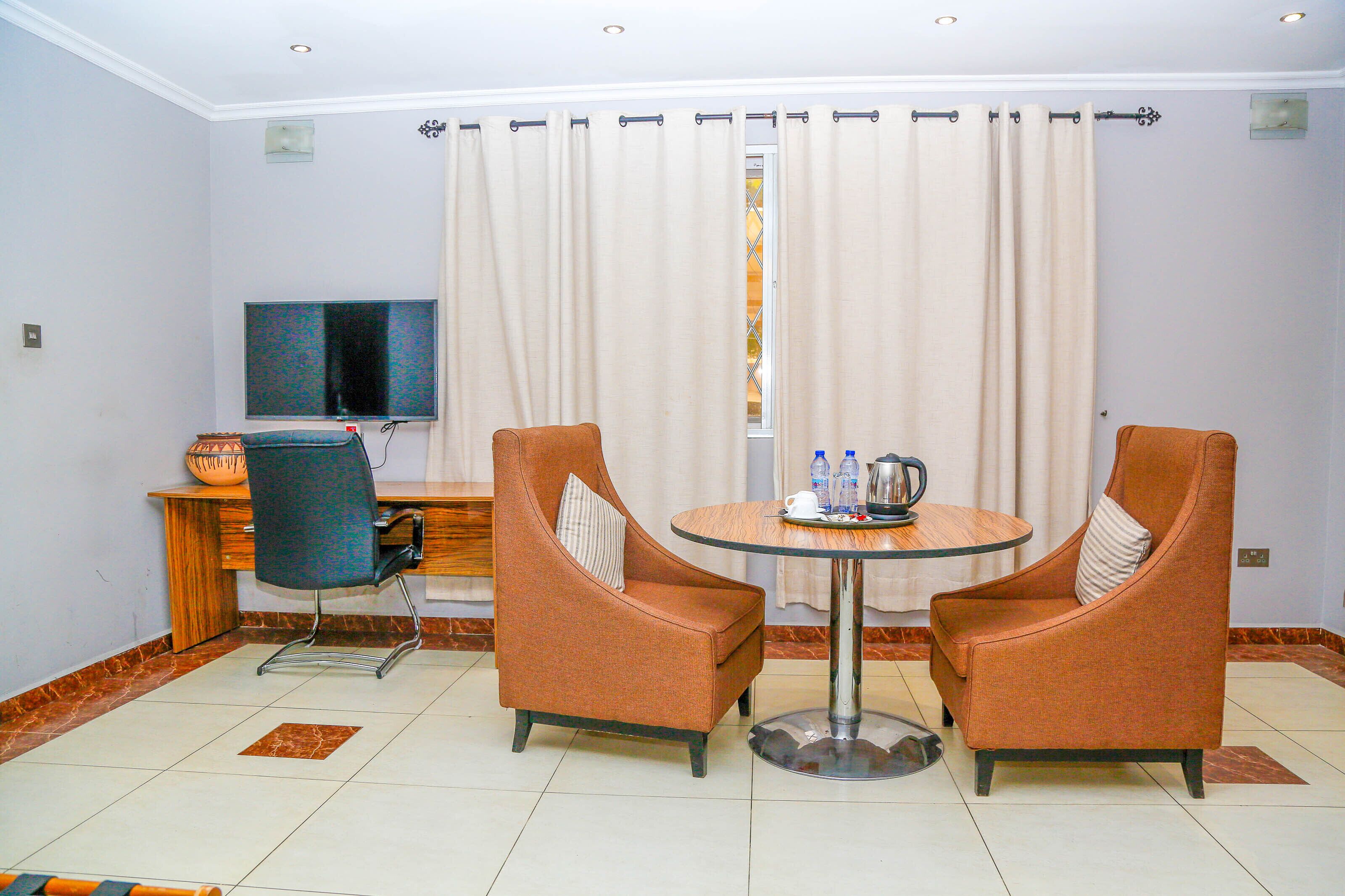 A hotel room with a TV, two armchairs, a small table with bottles, and a desk with a bowl.