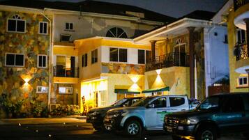 A multi-story hotel with a stone facade, multiple balconies, and a parking area with several cars.