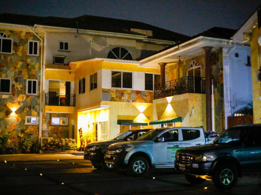 A multi-story hotel with a stone facade, multiple balconies, and a parking area with several cars.