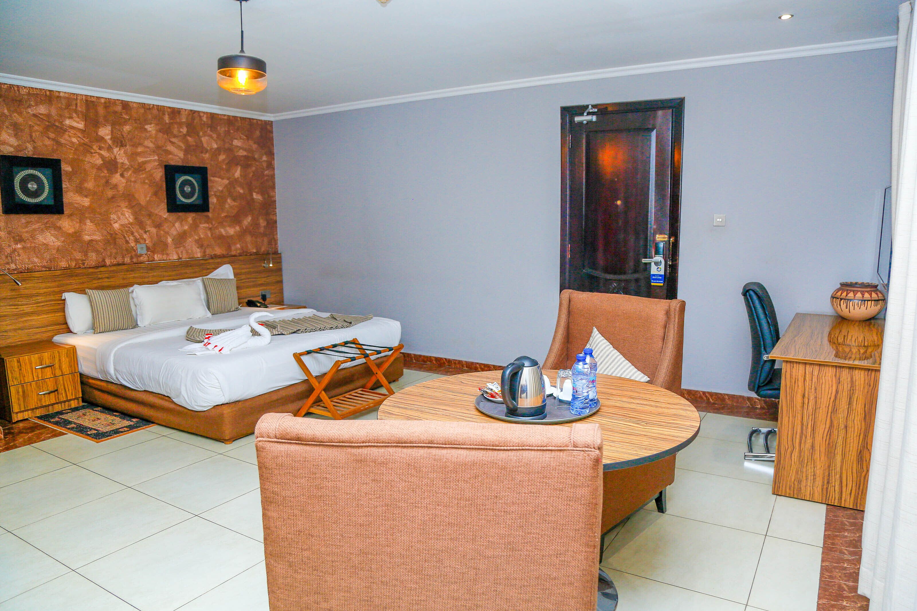 A hotel room with a bed, a chair, a small table with a kettle and water bottles, a wooden nightstand, and a door with a mirror.