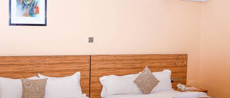 Une chambre d’hôtel avec une tête de lit en bois, deux lits avec du linge de lit blanc, des coussins décoratifs à motifs, une œuvre d’art encadrée au mur et un appareil de climatisation.