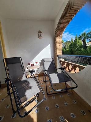 Ein Balkon mit zwei Loungestühlen, einem kleinen Tisch mit Getränken und Blick auf Grünflächen und Gebäude.
