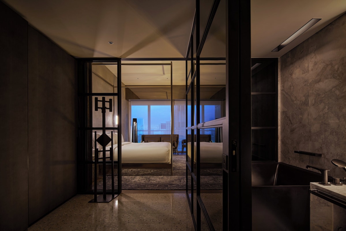 A modern hotel room with a glass partition, a bed, a desk, and a view of the cityscape.