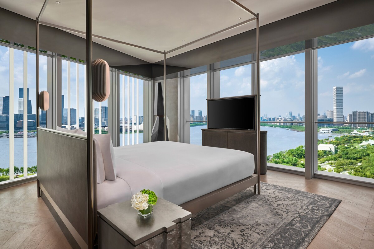 A modern hotel room with a large bed, a TV, and a view of a cityscape and water.