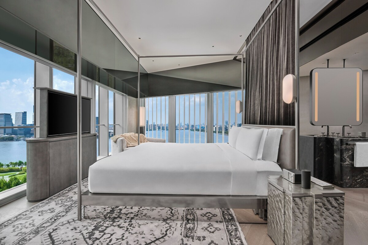 A modern bedroom with a large bed, a TV, and a view of a cityscape.