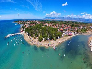 Vista aérea de un pueblo costero con playa, barcos y montañas al fondo.