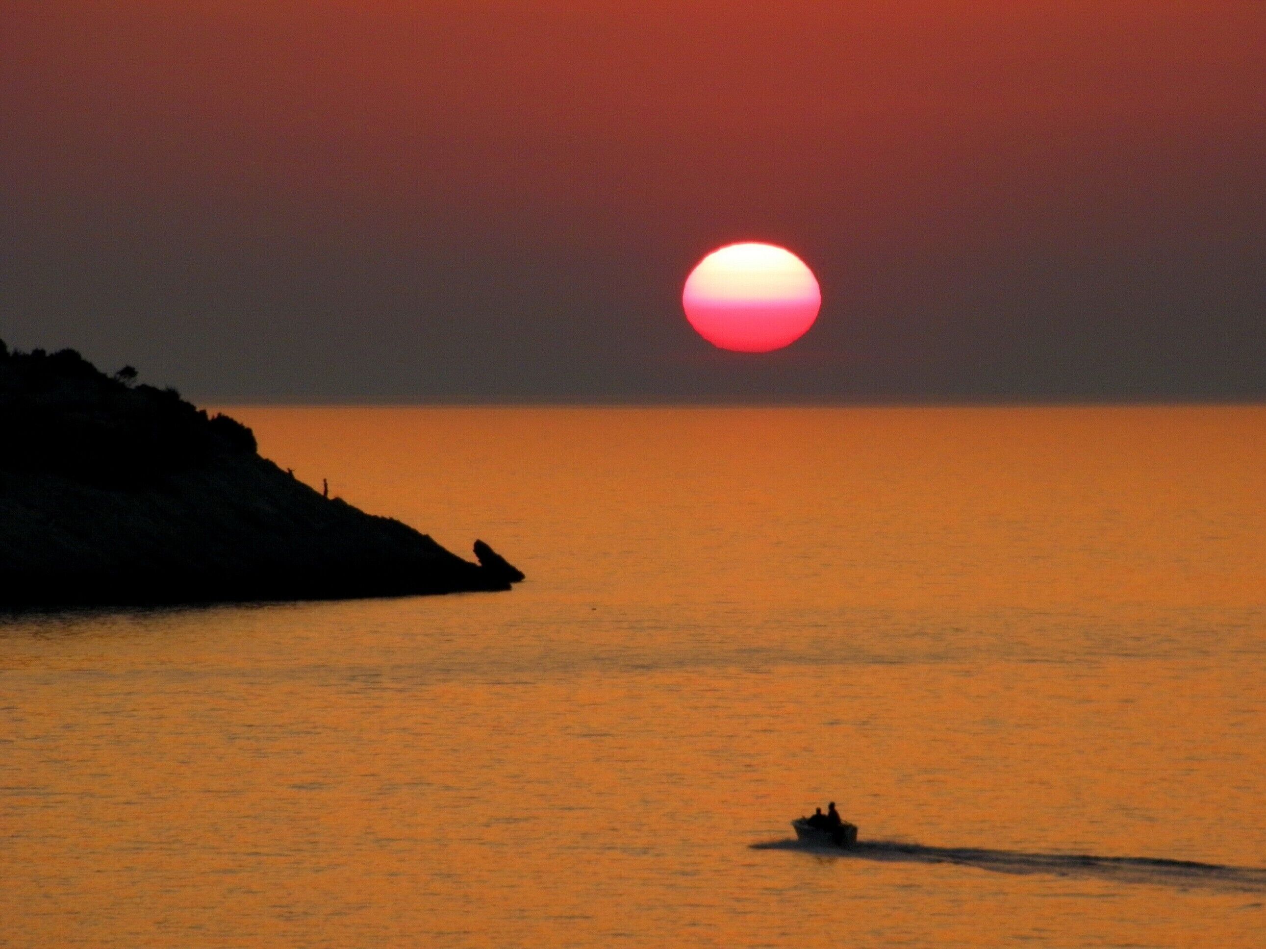 A sunset over a calm sea with a silhouette of a boat and a rocky coastline.