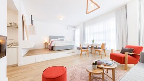 A modern hotel room with a bed, a dining area, a red armchair, and a small table with a book and a vase. - THE Premium Loft Apartment - 1-common Terrace (Piraeus)