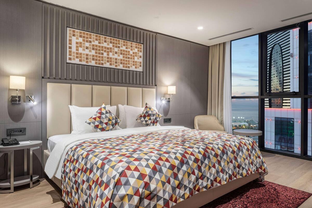 Ein modernes Hotelzimmer mit einem großen Bett, Blick auf die Stadt und einer gemusterten Bettdecke.