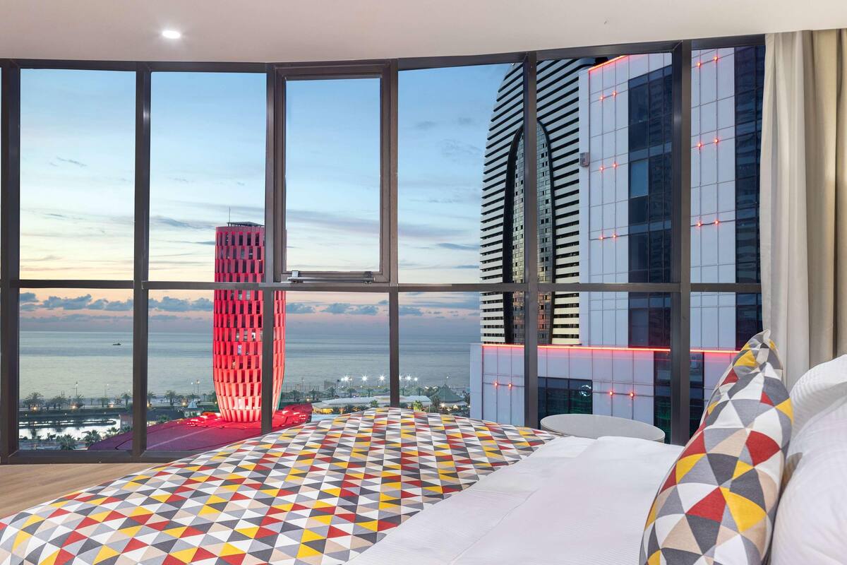 Ein Hotelzimmer mit großen Fenstern und Blick auf die Stadt, einem gemusterten Boden und einem Bett mit Kissen.