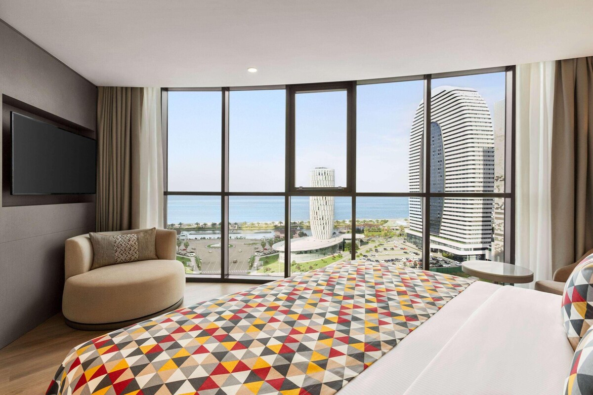Une chambre d’hôtel moderne dotée d’un grand lit, d’un canapé, d’une télévision et offrant une vue sur le paysage urbain et l’eau.