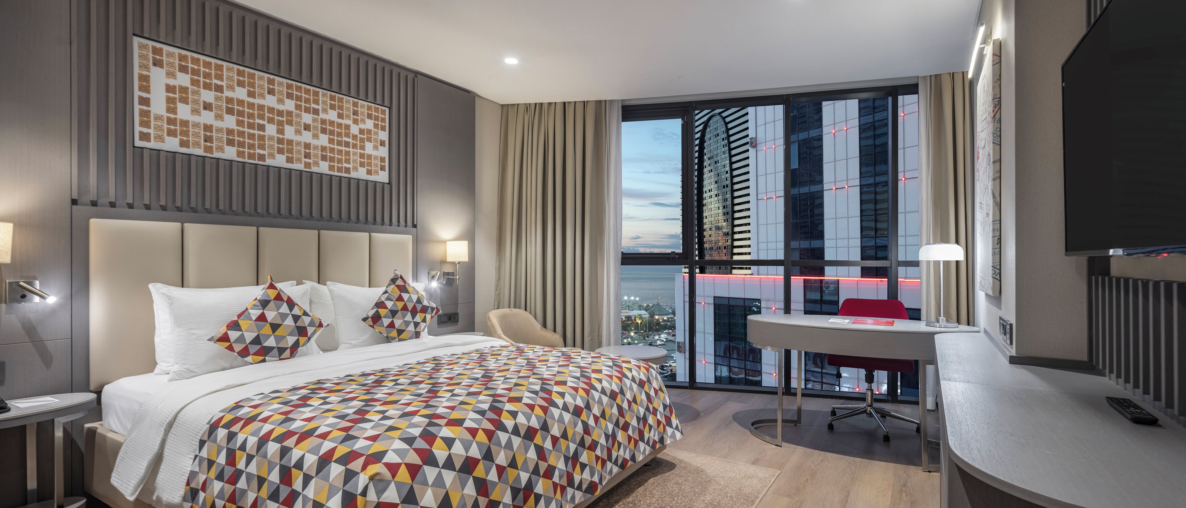 Een moderne hotelkamer met een groot bed, een bureau en uitzicht op een stadsgezicht door middel van kamerhoge ramen.
