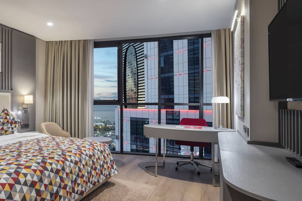 Ein modernes Hotelzimmer mit einem großen Bett, einem Schreibtisch und Blick auf die Skyline durch raumhohe Fenster.