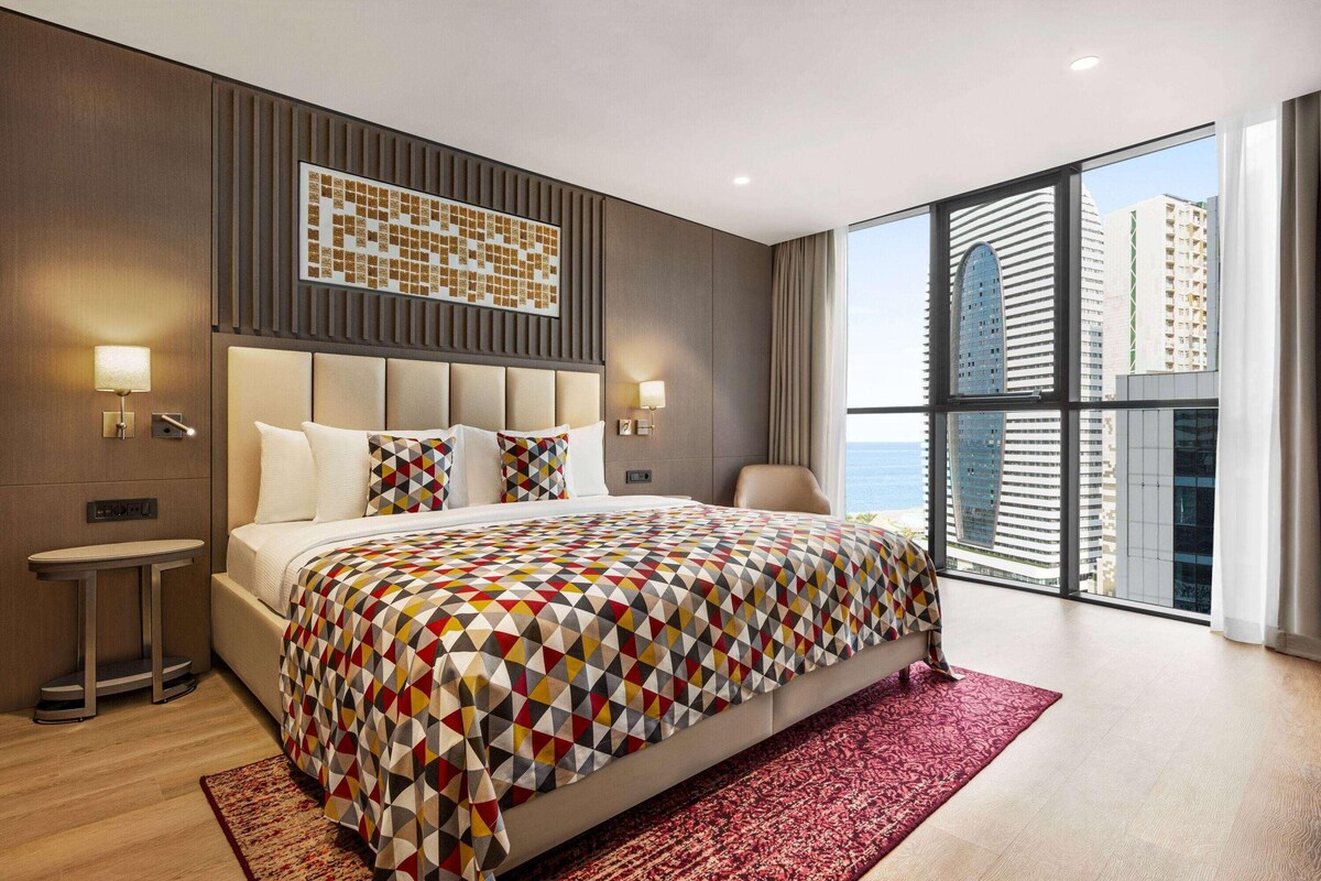 Une chambre d’hôtel moderne avec un grand lit, une petite table de chevet, un tapis rouge et une vue sur la ville à travers de grandes fenêtres.