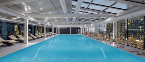 An indoor swimming pool with a skylight ceiling, exercise equipment, and lounge chairs.