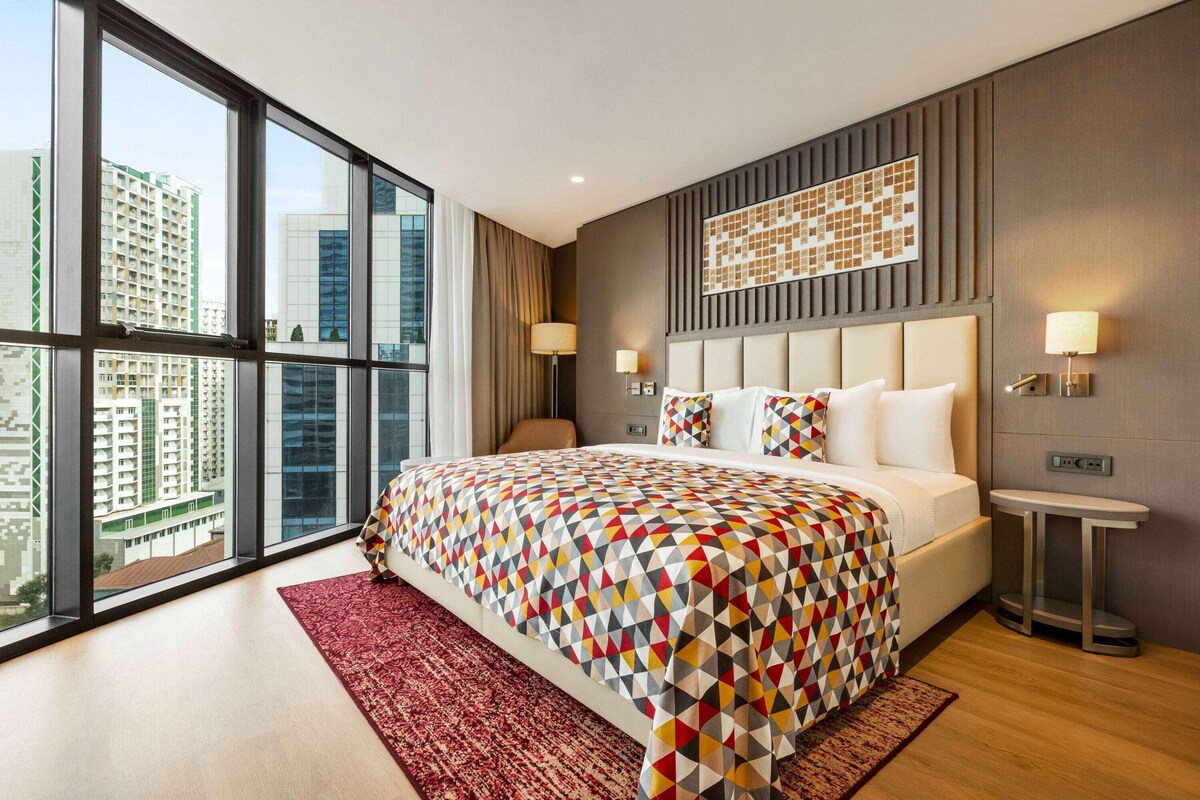 Une chambre d’hôtel moderne avec un grand lit, une vue sur la ville grâce à de grandes fenêtres du sol au plafond, un tapis rouge et une petite table de chevet.