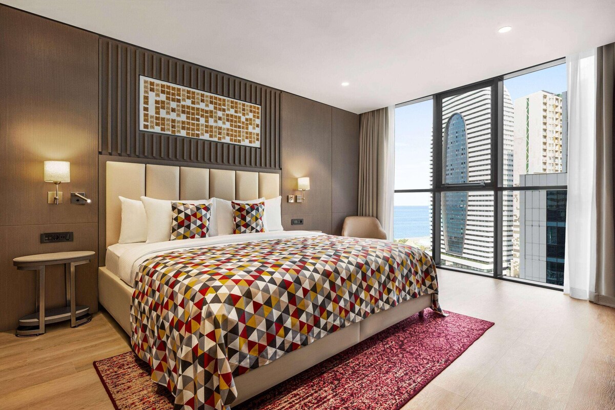 Une chambre d’hôtel moderne avec un grand lit, une petite table de chevet, un tapis rouge et une vue sur la ville à travers de grandes fenêtres.