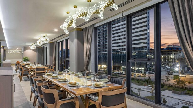 Een moderne eethoek met een lange houten tafel gedekt voor een maaltijd, grote ramen met uitzicht op de stad en decoratieve verlichting.