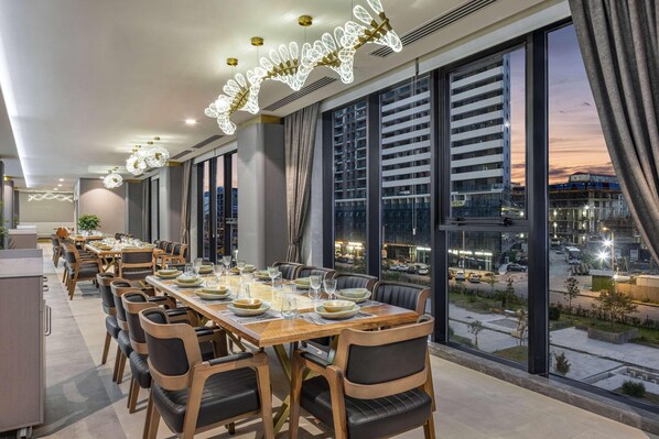 Un espace de restauration moderne avec une longue table en bois dressée pour un repas, de grandes fenêtres offrant une vue sur la ville et un éclairage décoratif.