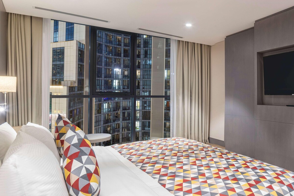 Une chambre d’hôtel moderne avec un grand lit, une télévision, une fenêtre donnant sur la ville et une petite table.