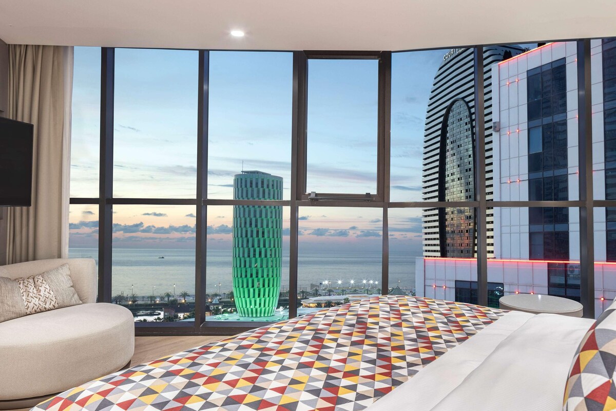 Une chambre d’hôtel moderne dotée d’une grande fenêtre donnant sur la ville, d’un lit recouvert d’une couvre-lit à motifs géométriques et d’un coin salon avec un canapé et une table ronde.