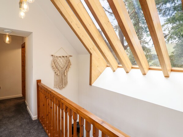 A room with a sloped ceiling, wooden beams, a macrame wall hanging, and a view of trees through a large window. - Ysgubor Craig Y Gronfa (Machynlleth)