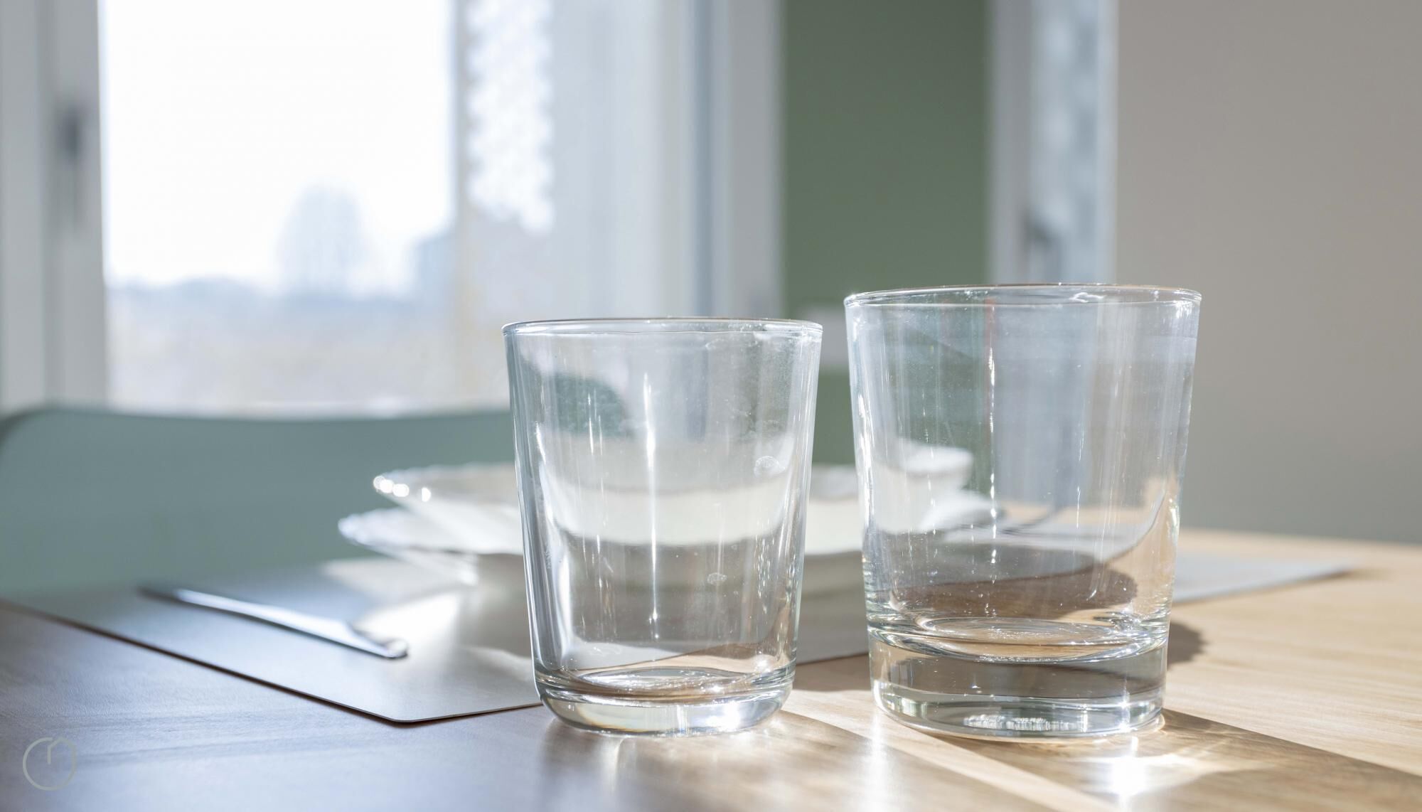 Two clear glasses on a wooden table with a napkin and a placemat, near a window with a view of buildings.
