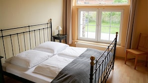 Un dormitorio con cama, mesita de noche, una silla y ventana con vistas a la vegetación.