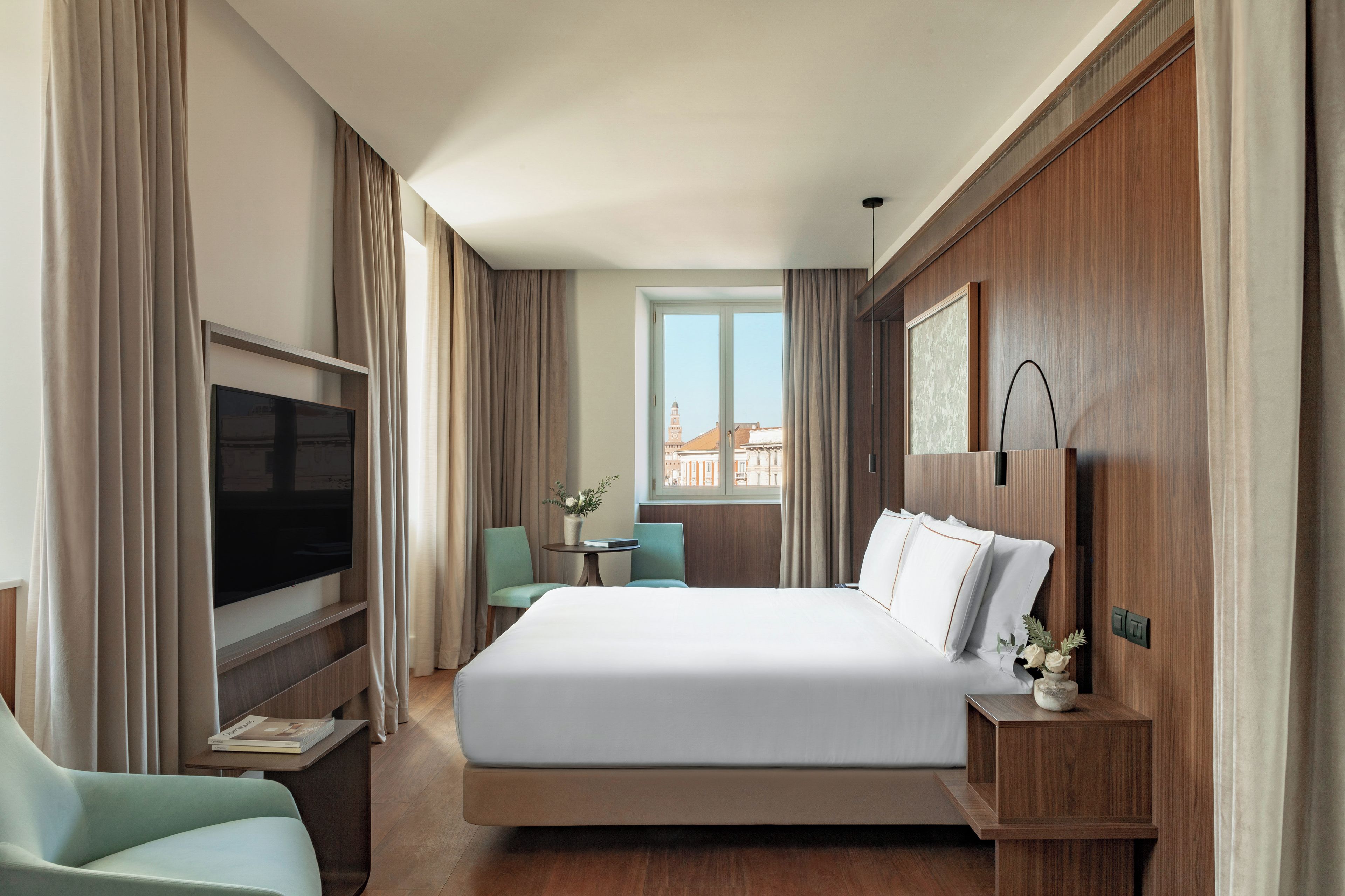 A hotel room with a large bed, a TV, a small table with a vase, and a view of buildings through the window.