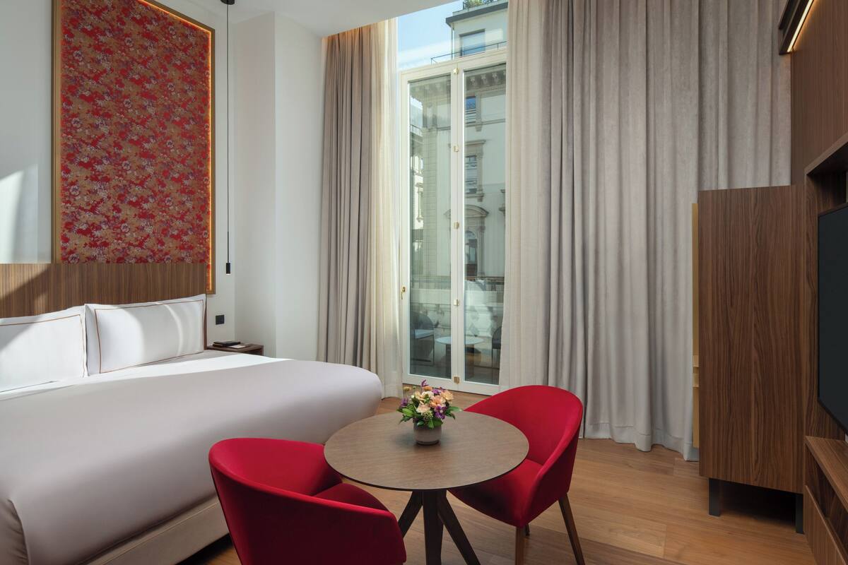 Une chambre d’hôtel avec un lit, une table ronde entourée de chaises, un téléviseur et une vue sur l’extérieur, visible à travers une porte vitrée.