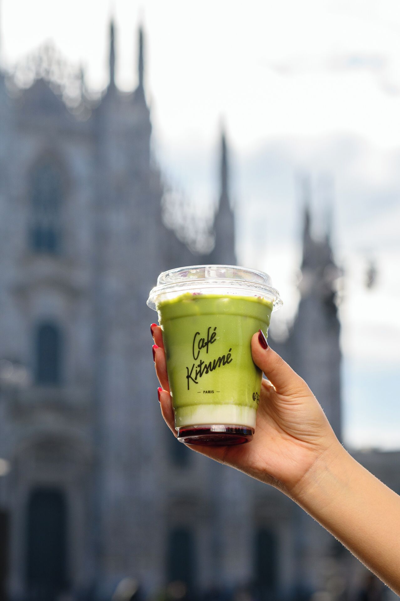 Una mano sostiene una taza con bebida verde que lleva la inscripción 'Café Kitsuné Paris', sobre un fondo borroso de un edificio con dos torres.