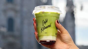 Una mano sostiene una taza con bebida verde que lleva la inscripción 'Café Kitsuné Paris', sobre un fondo borroso de un edificio con dos torres.