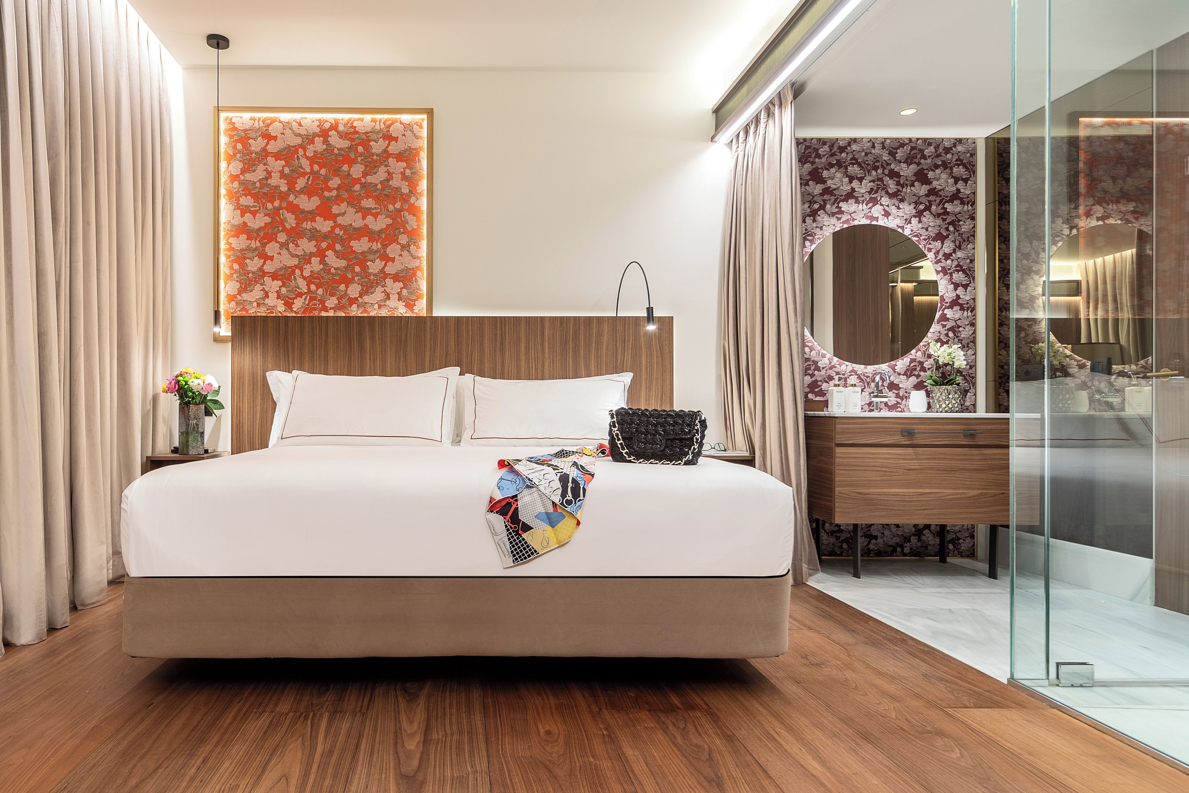 Une chambre d’hôtel moderne dotée d’un grand lit, d’une tête de lit en bois, d’une cabine de douche en verre et d’une salle de bain avec un miroir rond et un meuble-lavabo en bois.