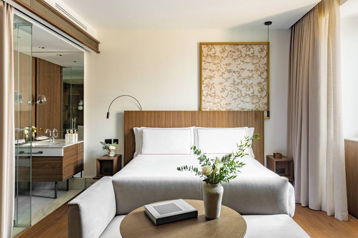 Une chambre d’hôtel moderne dotée d’un grand lit, d’une table de chevet avec une lampe, d’une salle de bain fermée par des parois de verre et d’un élément décoratif mural.