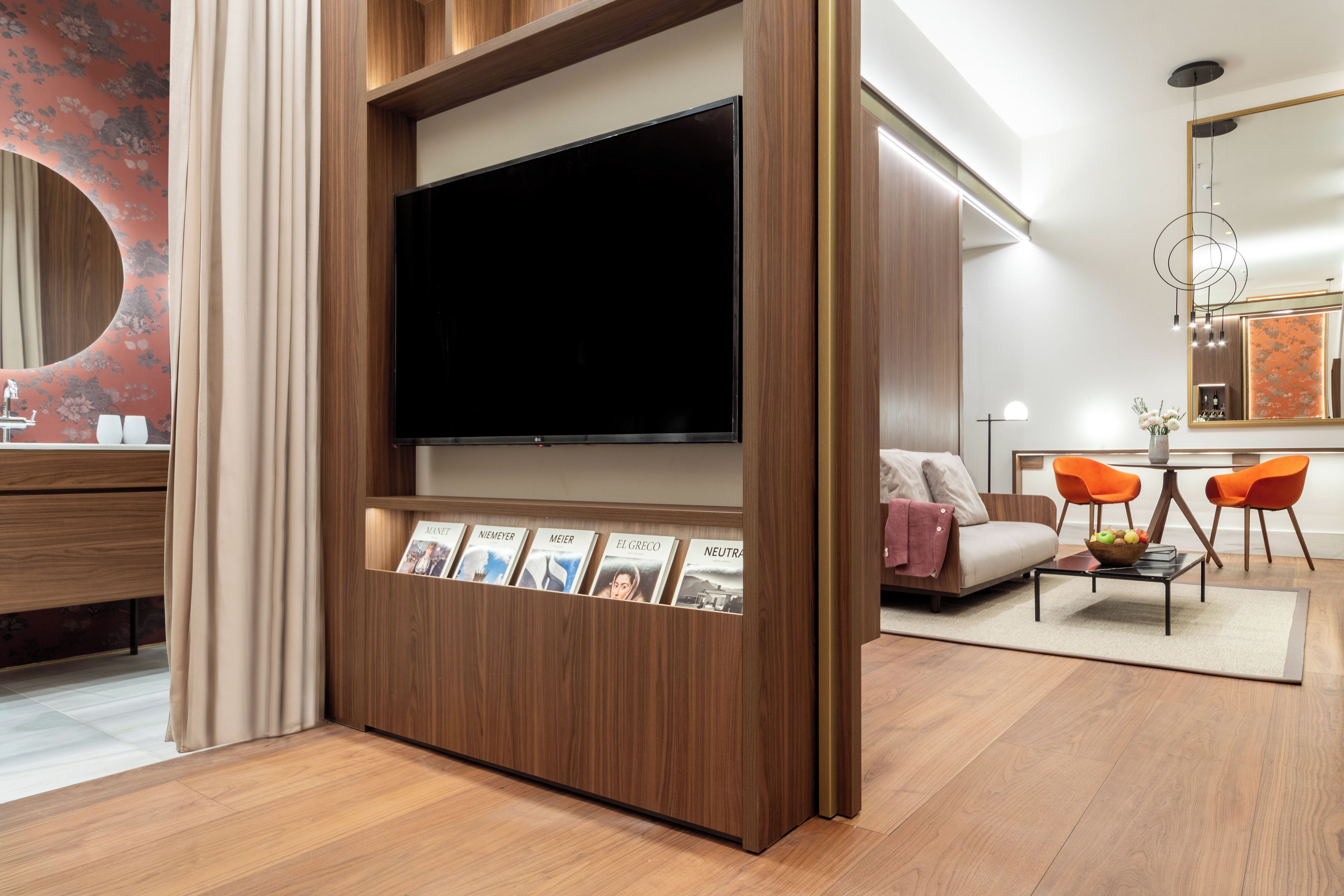 A modern living room with a large flat-screen TV, wooden shelving with magazines, a sofa, two orange chairs, and a dining table.