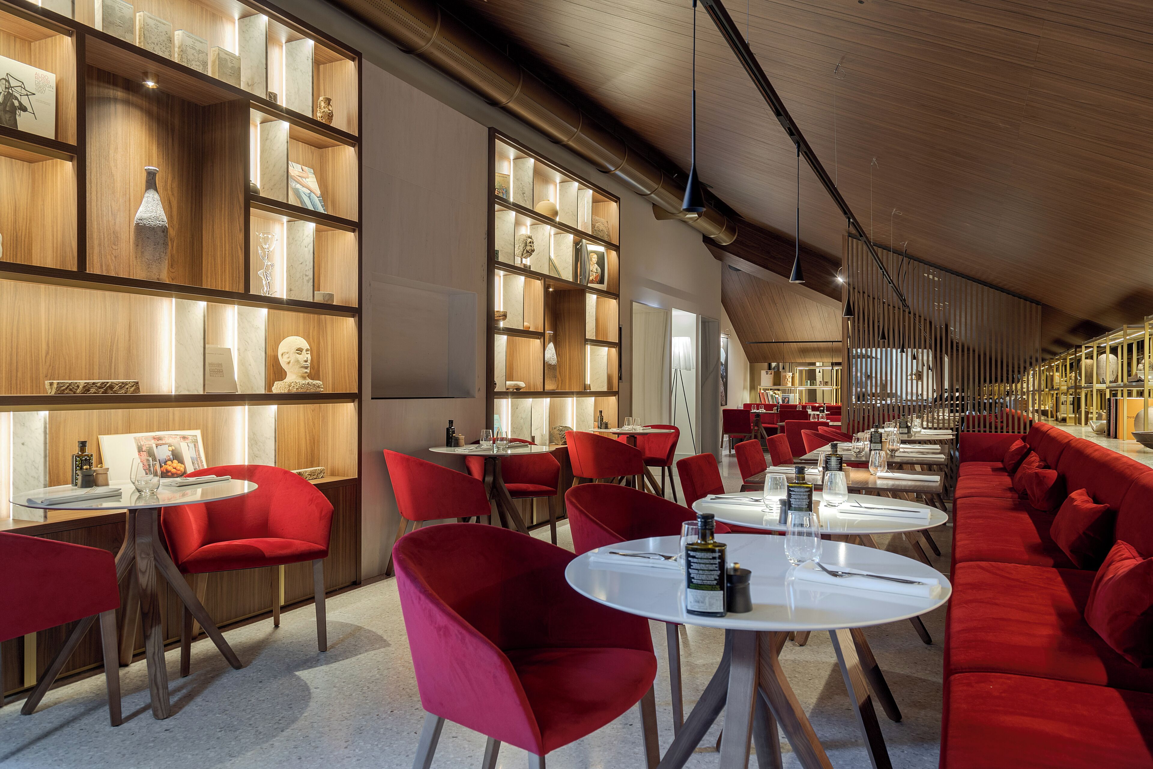 A modern restaurant with red chairs and white tables, wooden shelves with decorative items, and a sloped ceiling with exposed beams.
