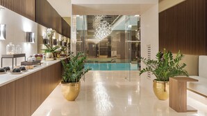 Un hall d’hôtel moderne avec un comptoir d’accueil, des plantes en pot, un espace piscine et un lustre.