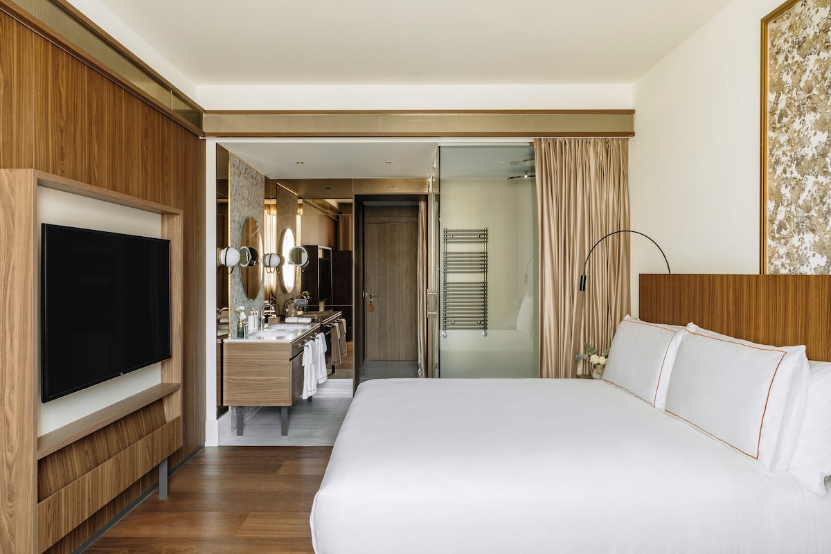 A hotel room with a large bed, a TV, a vanity with a mirror, and a shower area.