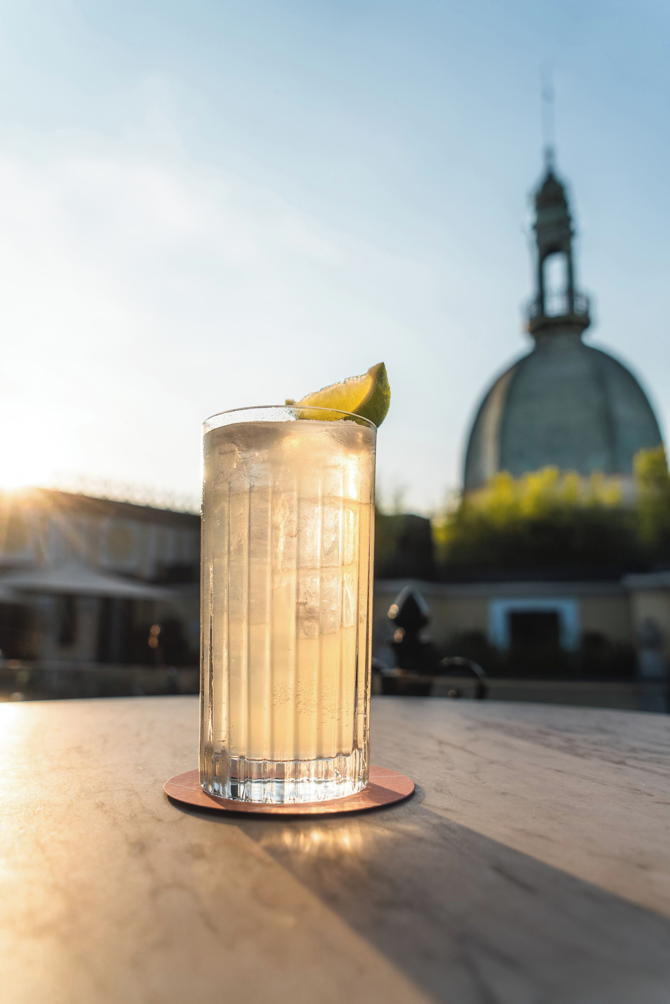 A clear glass with a lime wedge on a coaster, placed on a table with a building in the background.