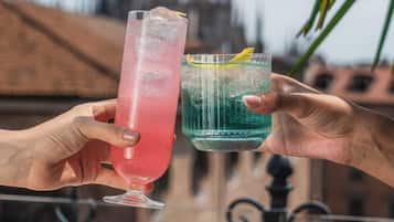 Two hands holding cocktail glasses, a table with food, and a building in the background.