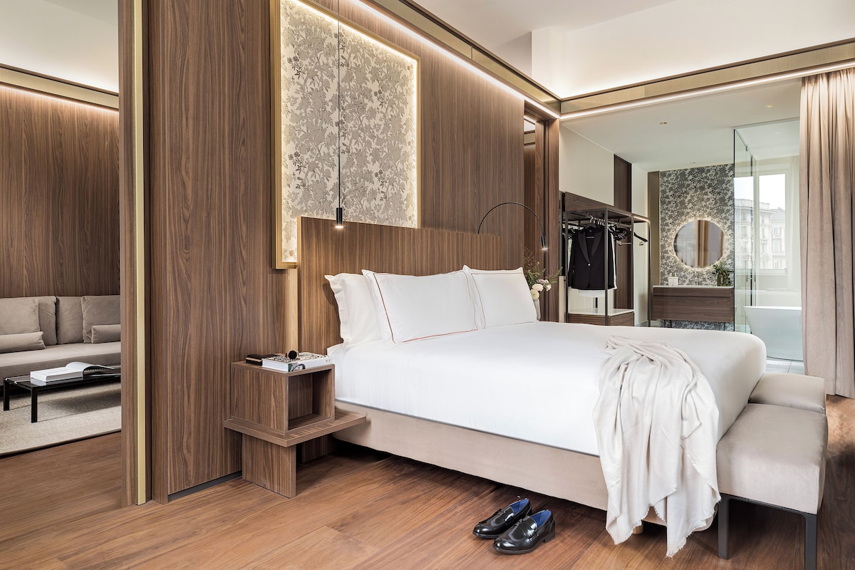 A modern bedroom with a large bed, wooden paneling, a nightstand, a mirror, and a view of the city through a glass door.