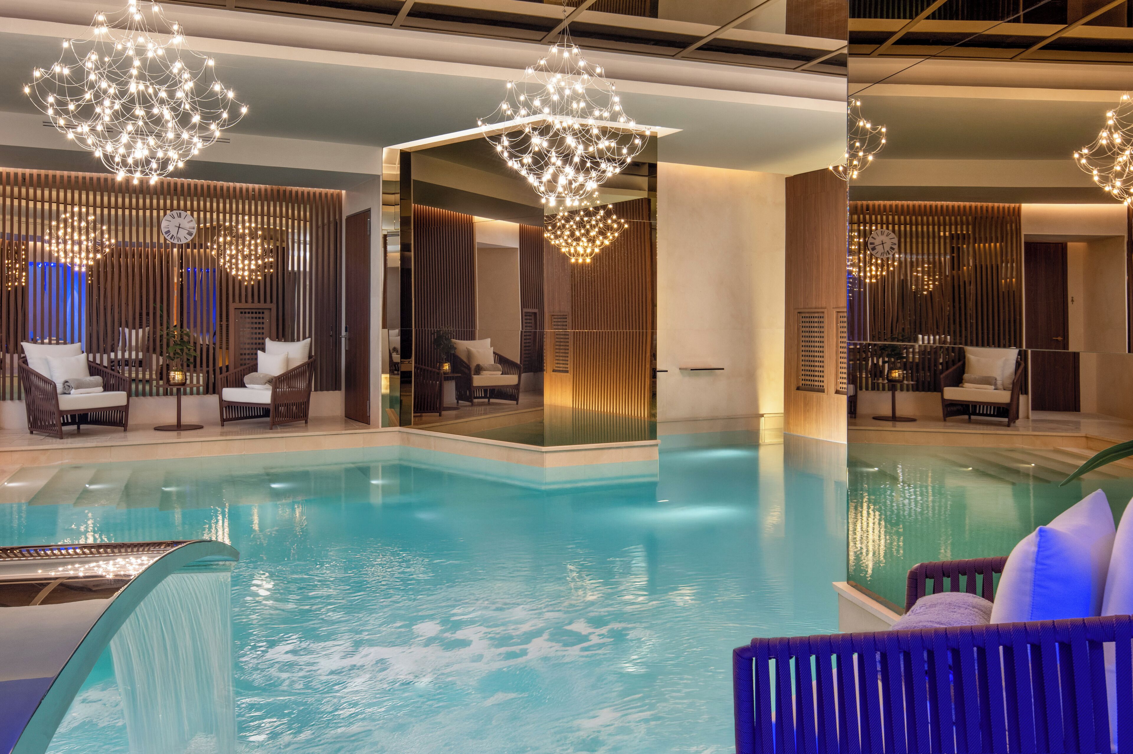 A modern indoor pool area with a glass barrier, lounge chairs, and decorative lighting.