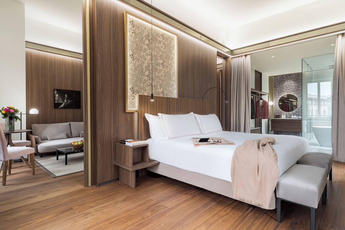 Une chambre d’hôtel moderne dotée d’un grand lit, d’un coin salon avec un canapé et un fauteuil, d’une petite table ornée d’un vase de fleurs et d’un grand miroir qui reflète la pièce.