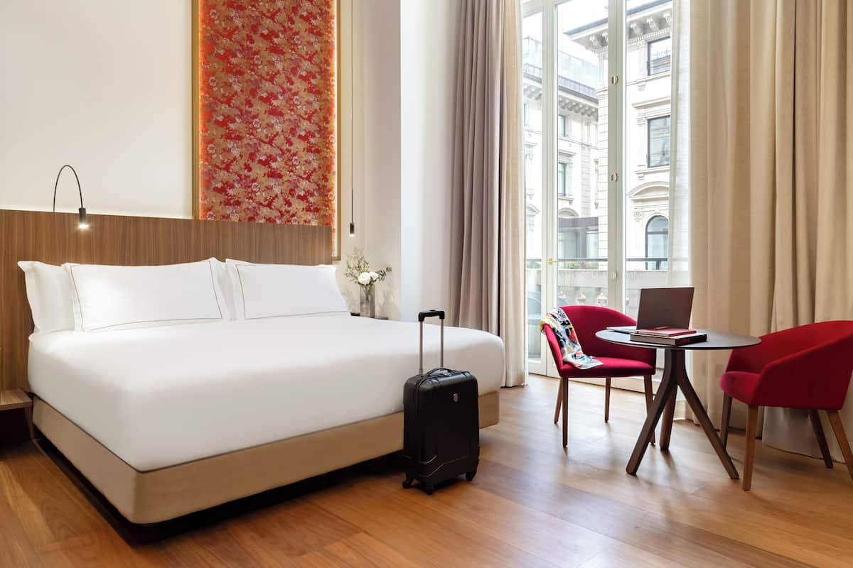 Une chambre d’hôtel avec un lit, une valise, un bureau avec un ordinateur portable et une vue sur la ville grâce à de grandes fenêtres.