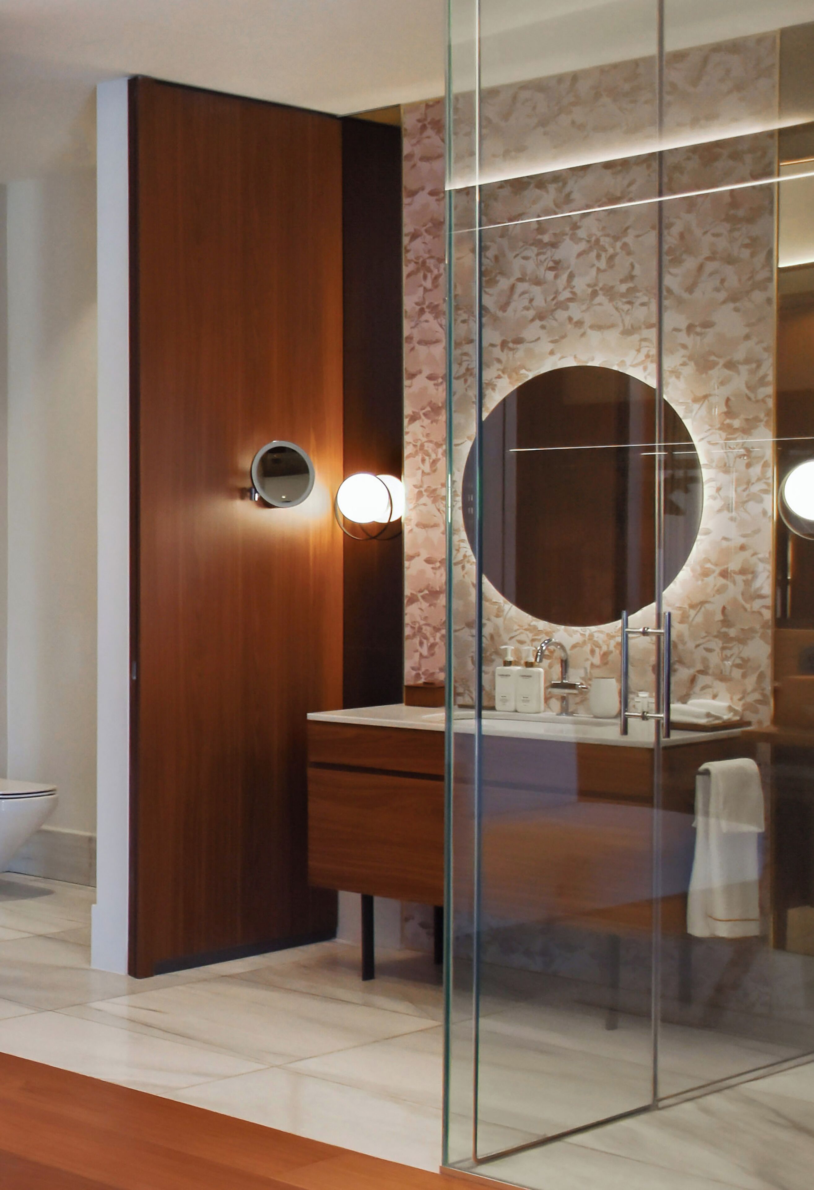 A modern bathroom with a wooden door, a glass partition, a round mirror, and a vanity with a sink.