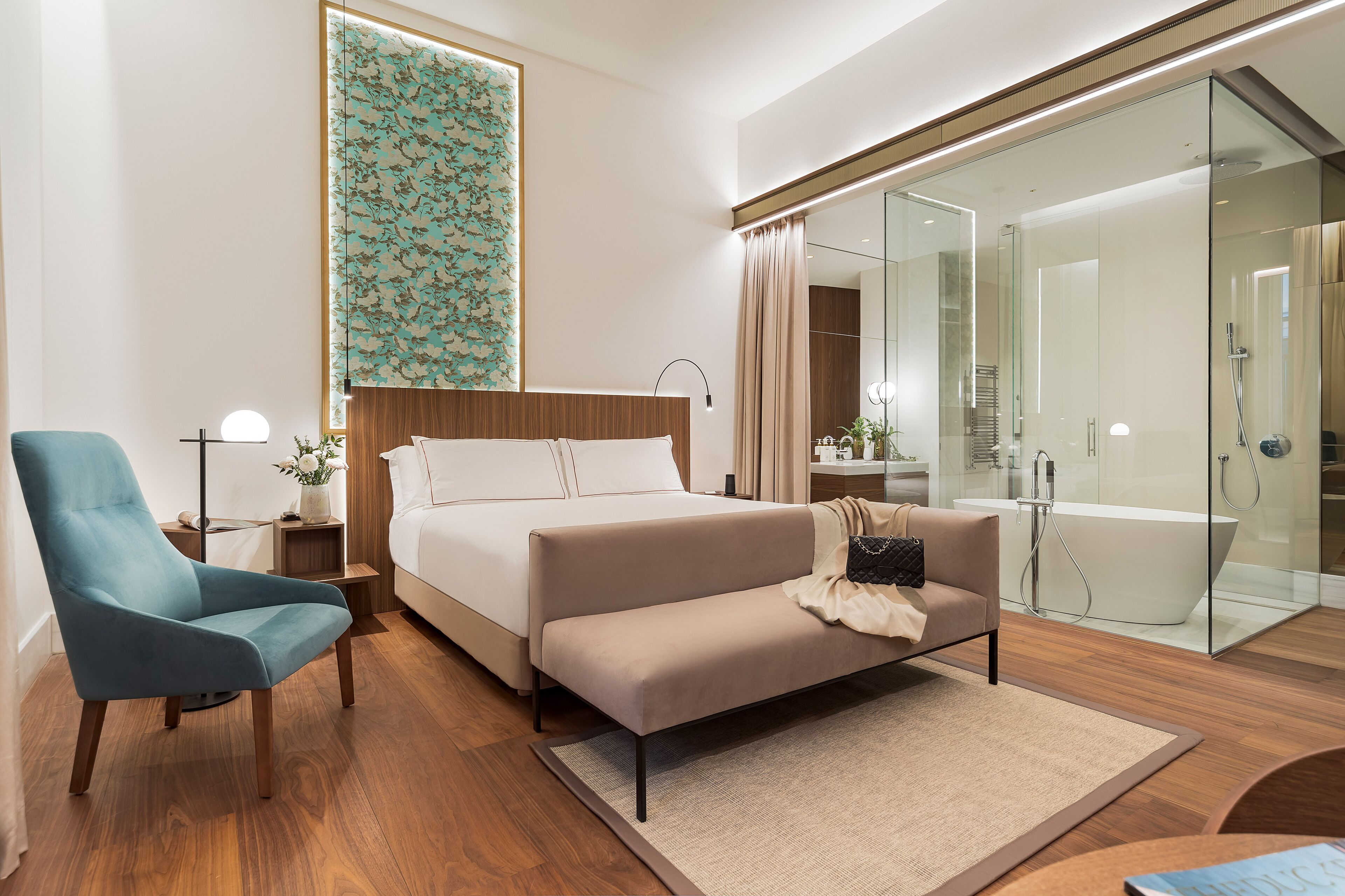 A modern hotel room with a bed, a sofa, a chair, a glass-enclosed shower, and a large window with a floral wallpaper.