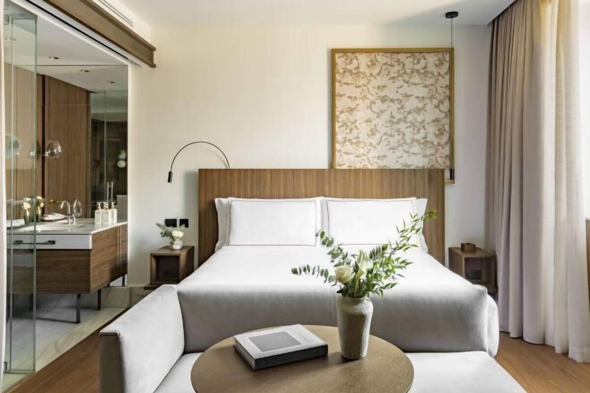 A modern hotel room with a large bed, a bedside table with a book, a vase with flowers, a mirror, and a wall-mounted artwork.