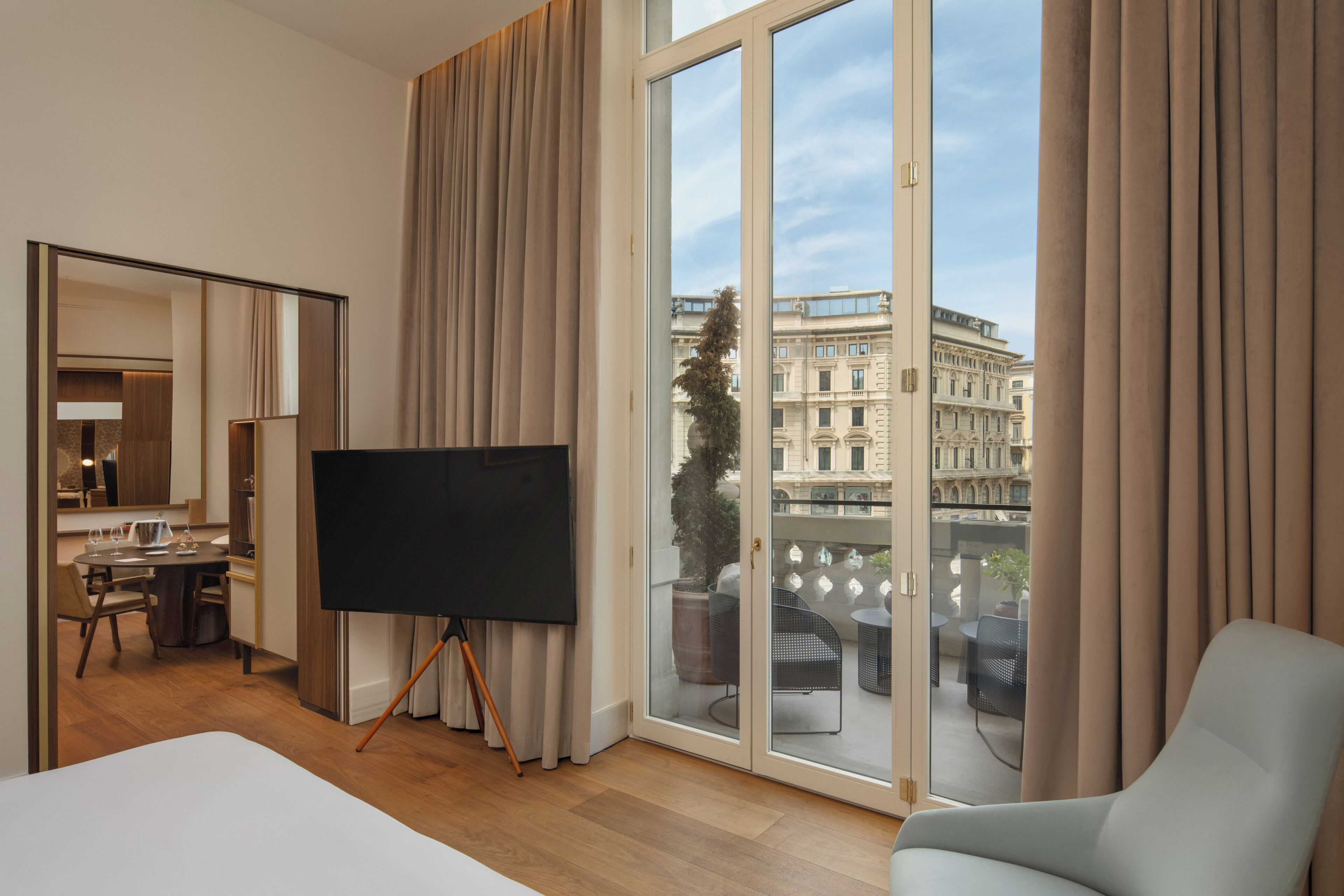 A hotel room with a large window, a flat-screen TV, a dining area, and a balcony with outdoor furniture.
