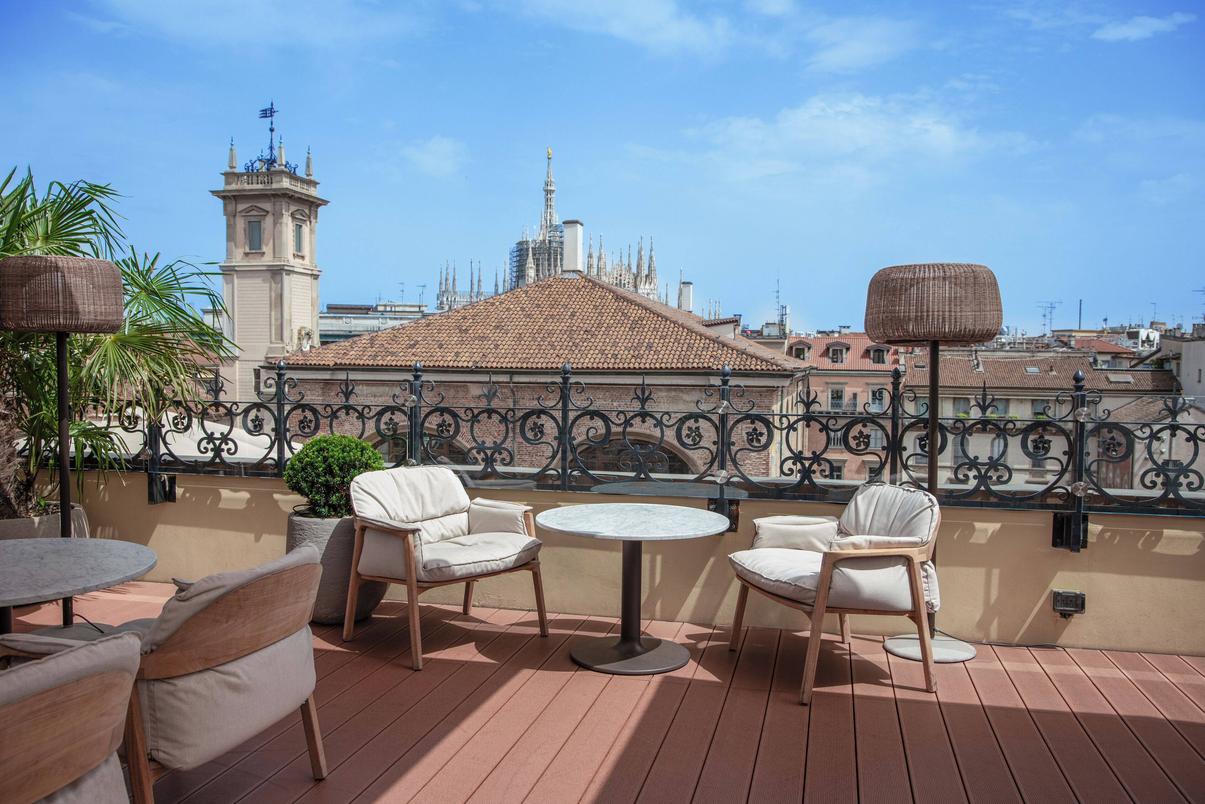 A terrace with wicker tables and chairs, a view of a historic building with a tower and spires, and a wrought iron railing.